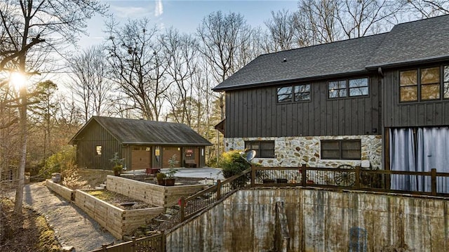 exterior space featuring a garage, a garden, a shingled roof, stone siding, and an outbuilding