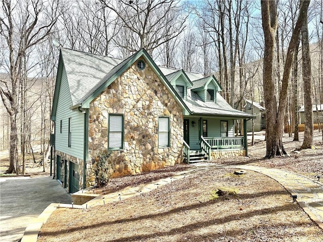 view of front of house featuring driveway, an attached garage, covered porch, a shingled roof, and stone siding