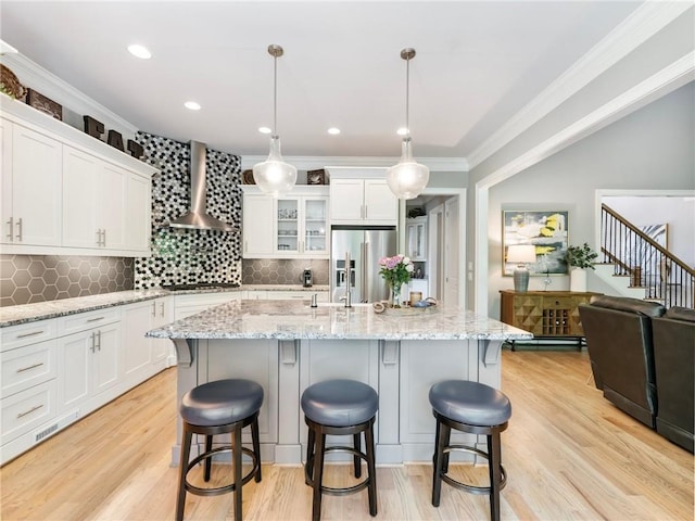 kitchen with hanging light fixtures, high quality fridge, white cabinets, a center island with sink, and wall chimney exhaust hood