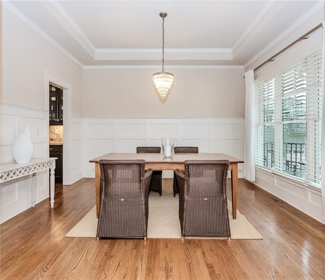 unfurnished dining area featuring a decorative wall, wood finished floors, a chandelier, a raised ceiling, and ornamental molding