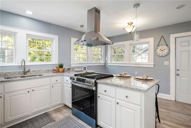 kitchen with stainless steel gas range oven, island range hood, hanging light fixtures, white cabinetry, and a sink