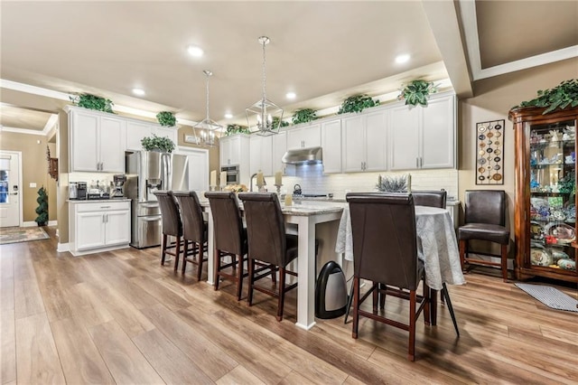 kitchen featuring light wood-type flooring, a center island with sink, stainless steel appliances, decorative light fixtures, and white cabinets