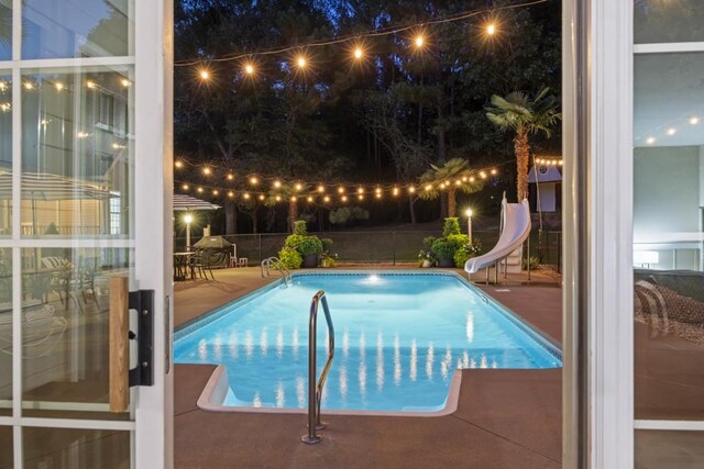 pool at night with a fenced in pool, a water slide, fence, and a patio area