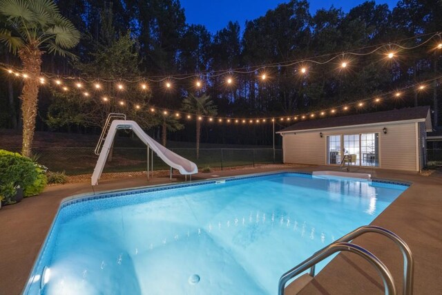 pool at night with an exterior structure, a water slide, fence, an outdoor structure, and an outdoor pool