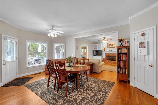 dining space featuring crown molding, a fireplace, baseboards, a ceiling fan, and light wood-style flooring