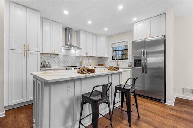 kitchen with stainless steel refrigerator with ice dispenser, dark wood finished floors, a center island, white cabinetry, and wall chimney range hood