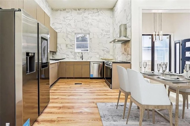 kitchen featuring pendant lighting, light hardwood / wood-style flooring, stainless steel appliances, decorative backsplash, and wall chimney exhaust hood