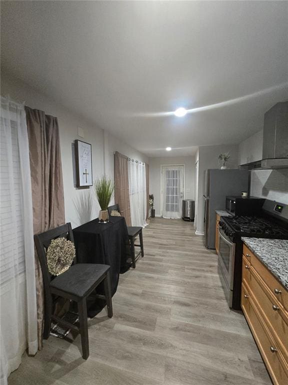 kitchen with gas range, light wood-style flooring, wall chimney exhaust hood, brown cabinets, and light stone countertops