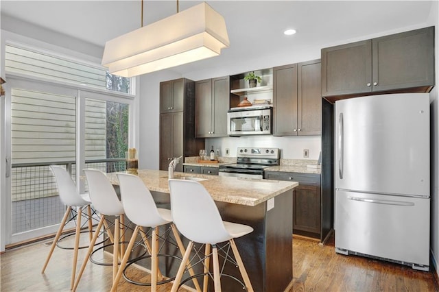 kitchen featuring an island with sink, wood finished floors, stainless steel appliances, a kitchen bar, and open shelves