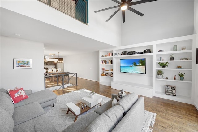 living room featuring ceiling fan with notable chandelier, built in shelves, wood finished floors, and a towering ceiling