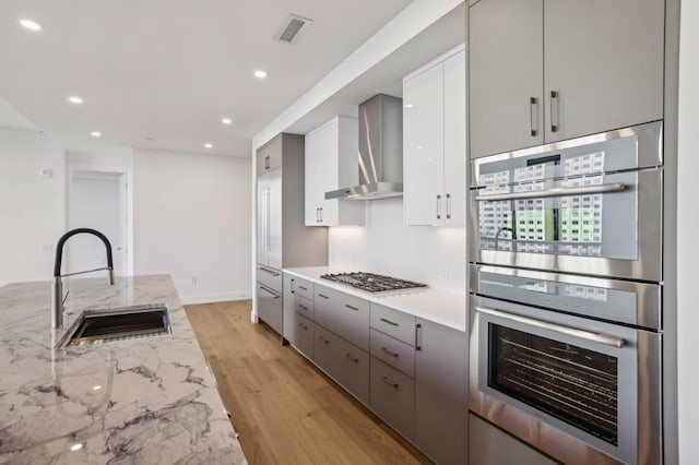 kitchen with wall chimney range hood, sink, light wood-type flooring, appliances with stainless steel finishes, and light stone counters