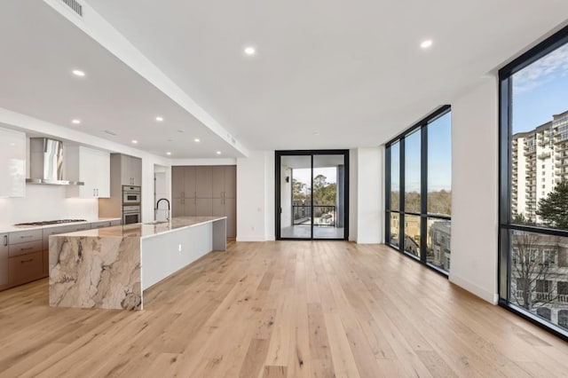 kitchen featuring floor to ceiling windows, a large island with sink, white cabinets, wall chimney exhaust hood, and light wood-type flooring