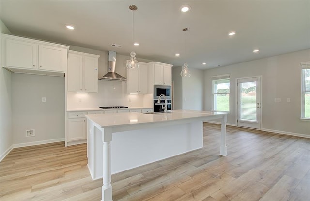 kitchen with light hardwood / wood-style floors, stainless steel microwave, wall chimney range hood, and an island with sink