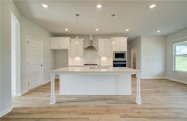 kitchen with light hardwood / wood-style flooring, oven, stainless steel microwave, a kitchen island with sink, and wall chimney range hood