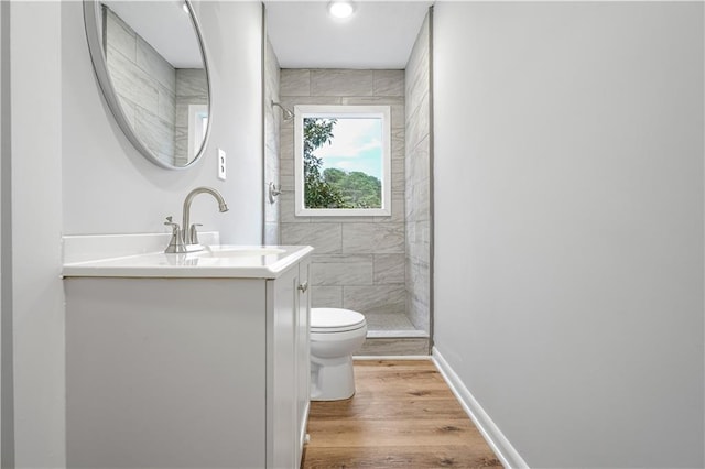 bathroom featuring toilet, vanity, wood finished floors, tiled shower, and baseboards