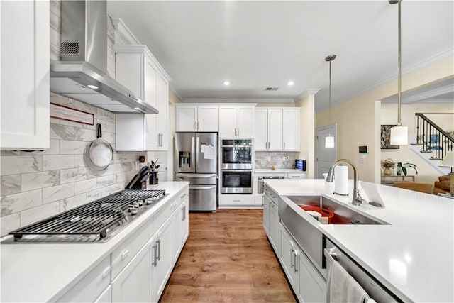 kitchen with appliances with stainless steel finishes, wall chimney range hood, pendant lighting, light hardwood / wood-style floors, and white cabinetry