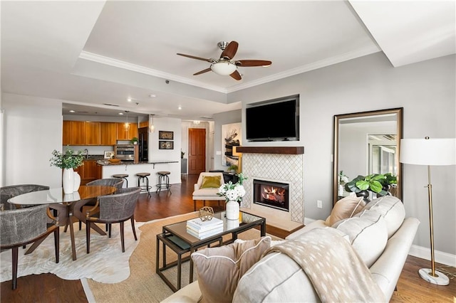 living area featuring baseboards, a tiled fireplace, ornamental molding, wood finished floors, and a ceiling fan