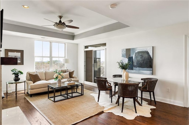 living area with a ceiling fan, a tray ceiling, dark wood finished floors, crown molding, and baseboards