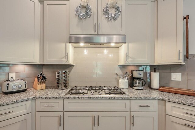 kitchen with stainless steel gas cooktop, under cabinet range hood, white cabinets, light stone counters, and backsplash