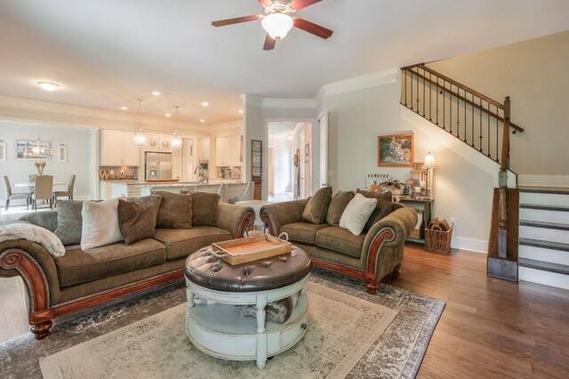 living room featuring crown molding, wood finished floors, stairway, baseboards, and ceiling fan with notable chandelier