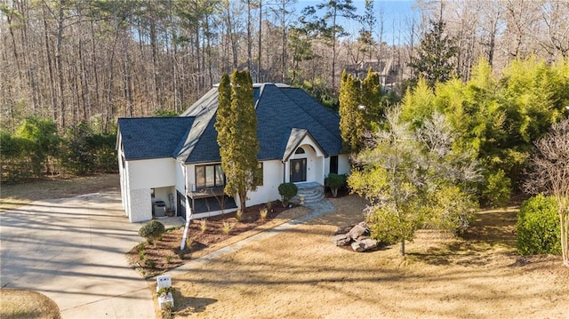 view of front of house with central AC unit, a shingled roof, concrete driveway, stucco siding, and a wooded view