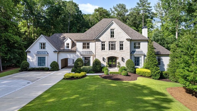 french country style house featuring a standing seam roof, a chimney, concrete driveway, a front yard, and stone siding