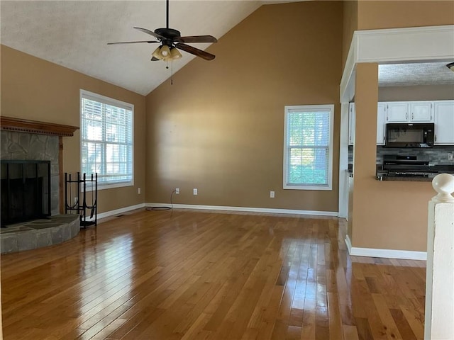 unfurnished living room featuring light wood-type flooring, a textured ceiling, high vaulted ceiling, a stone fireplace, and ceiling fan