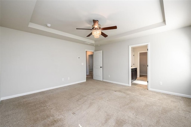 unfurnished bedroom with a tray ceiling, ensuite bathroom, light carpet, ceiling fan, and baseboards