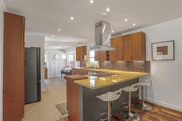 kitchen with light stone counters, brown cabinetry, island exhaust hood, freestanding refrigerator, and a sink