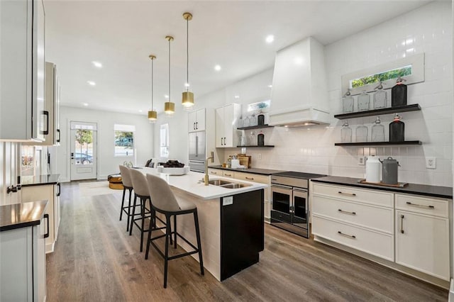 kitchen with dark wood-type flooring, a breakfast bar, custom range hood, a kitchen island with sink, and white cabinets