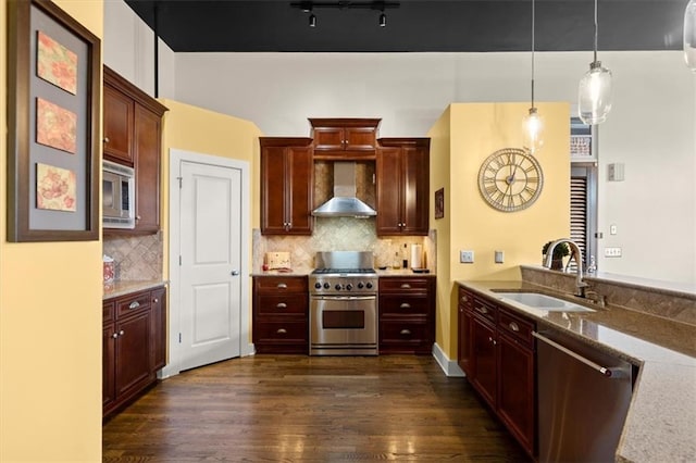 kitchen featuring dark wood finished floors, stainless steel appliances, hanging light fixtures, a sink, and wall chimney exhaust hood