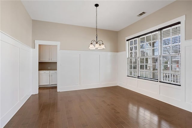unfurnished dining area with wainscoting, a decorative wall, dark wood-style floors, and an inviting chandelier
