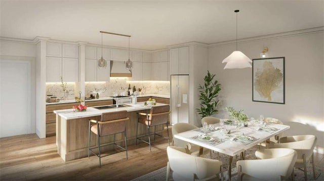 kitchen with white cabinetry, hanging light fixtures, a center island with sink, built in fridge, and custom range hood