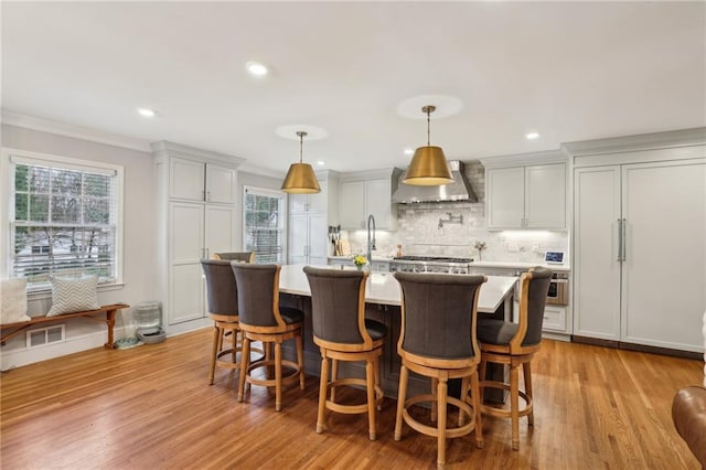 kitchen featuring crown molding, wall chimney range hood, a kitchen island with sink, and light wood finished floors