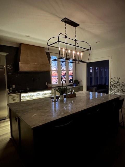 kitchen with decorative light fixtures, an island with sink, dark stone countertops, premium range hood, and stainless steel refrigerator