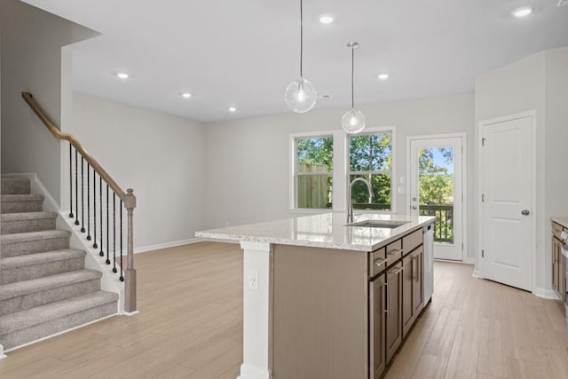 kitchen featuring an island with sink, light wood-type flooring, hanging light fixtures, light stone counters, and sink