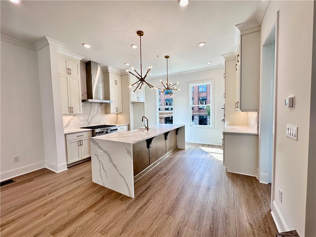 kitchen with a kitchen island with sink, wall chimney exhaust hood, hanging light fixtures, light wood-type flooring, and light stone counters