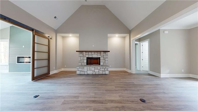 unfurnished living room with high vaulted ceiling, light hardwood / wood-style flooring, a stone fireplace, and a barn door