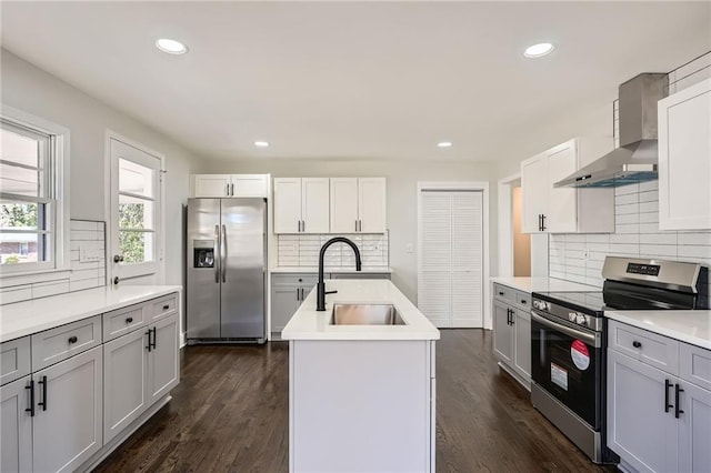 kitchen with sink, dark wood-type flooring, appliances with stainless steel finishes, a center island with sink, and wall chimney exhaust hood