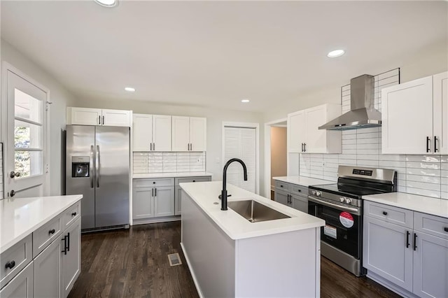 kitchen featuring wall chimney range hood, sink, appliances with stainless steel finishes, white cabinets, and a center island with sink