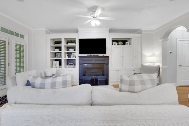 living room with arched walkways, a glass covered fireplace, ceiling fan, crown molding, and light wood-style floors