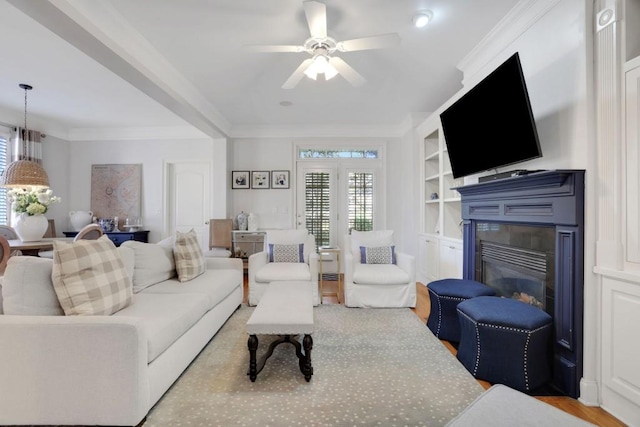 living area featuring built in shelves, wood finished floors, a ceiling fan, ornamental molding, and a glass covered fireplace