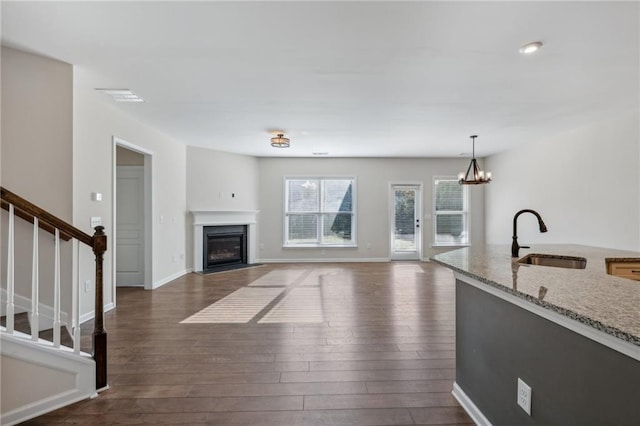 unfurnished living room with an inviting chandelier, a sink, stairway, dark wood-type flooring, and a glass covered fireplace