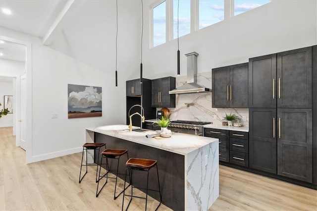 kitchen with wall chimney exhaust hood, light wood-type flooring, an island with sink, tasteful backsplash, and a kitchen bar