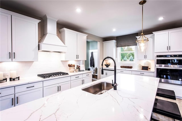 kitchen with appliances with stainless steel finishes, white cabinetry, sink, hanging light fixtures, and custom range hood