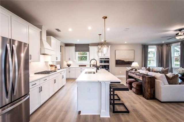 kitchen featuring premium range hood, hanging light fixtures, sink, stainless steel appliances, and a center island with sink