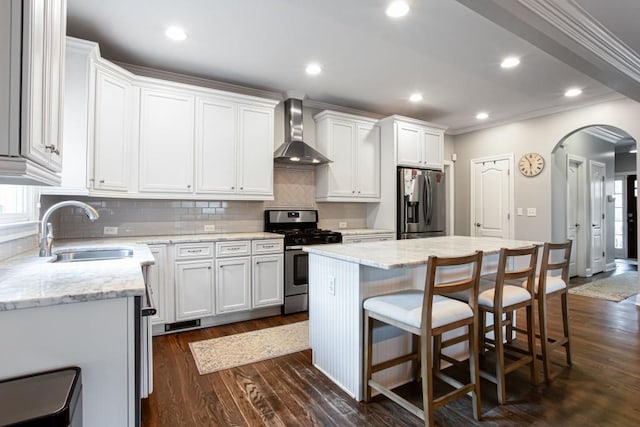 kitchen featuring arched walkways, appliances with stainless steel finishes, a center island, wall chimney range hood, and a sink