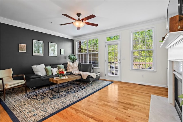 living room featuring baseboards, light wood-type flooring, a ceiling fan, crown molding, and a fireplace with flush hearth