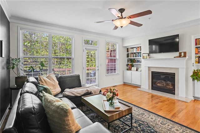 living room with ceiling fan, ornamental molding, a glass covered fireplace, wood finished floors, and built in shelves