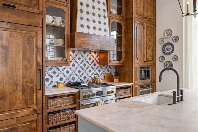 11277 Serenbe Ln, Chattahoochee Hills, GA 30268 - kitchen featuring a sink, stainless steel appliances, brown cabinetry, decorative backsplash, and glass insert cabinets